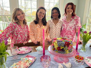Four women in pajamas standing around a table with a cake and candles, in a home setting.